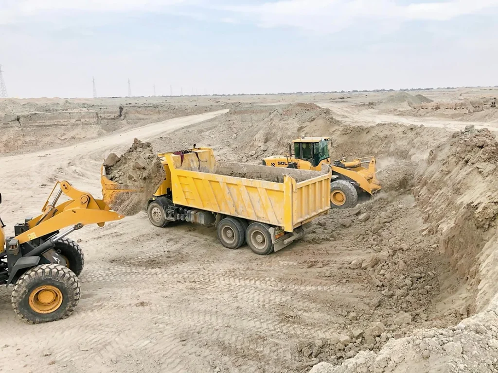 Yellow excavators load dirt into a yellow dump truck at a construction site in a barren, sandy landscape under a cloudy sky.