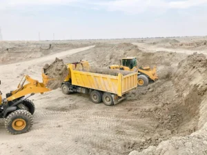 Yellow excavators load dirt into a yellow dump truck at a construction site in a barren, sandy landscape under a cloudy sky.