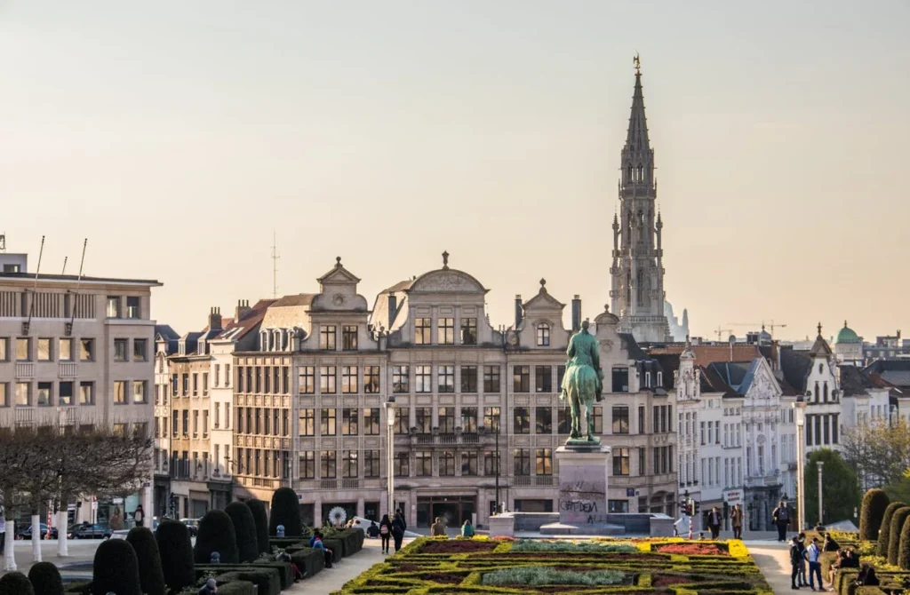 Cityscape featuring a bronze equestrian statue, gardens, and historic buildings under a clear sky. People walk along pathways, creating a tranquil mood.