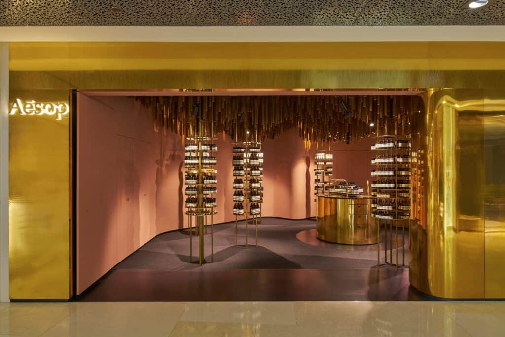 Brass-clad shopfront of Aesop ION in ION Orchard opening onto a pink interior beneath a canopy of hanging timber battens and slender brass display towers
