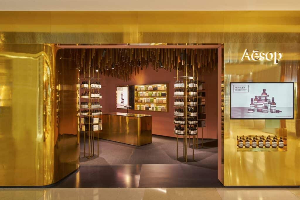 Front of Aesop ION with a curved polished brass wall, illuminated Aesop sign and screen beside an opening onto a brass counter and pink walls