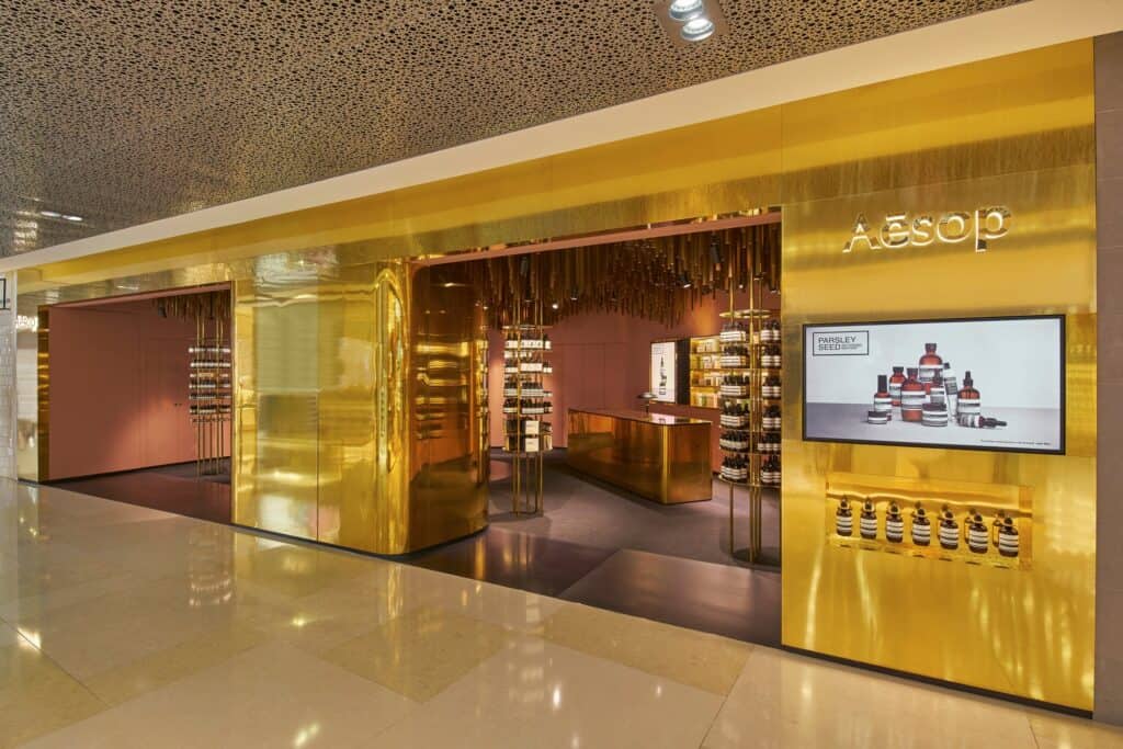 Long polished brass facade of Aesop ION along the marble-floored mall corridor of ION Orchard beneath a perforated metal ceiling
