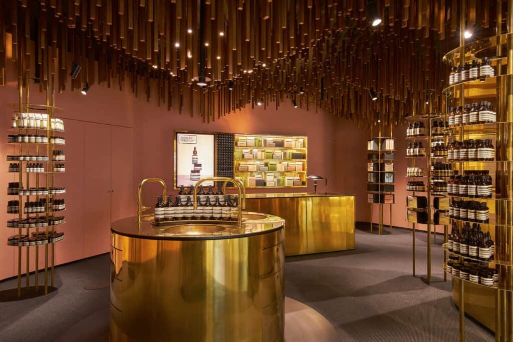 Cylindrical brass sink with brass swan-neck taps and amber bottles before a brass counter and lit product cabinet, timber battens hanging overhead at Aesop ION