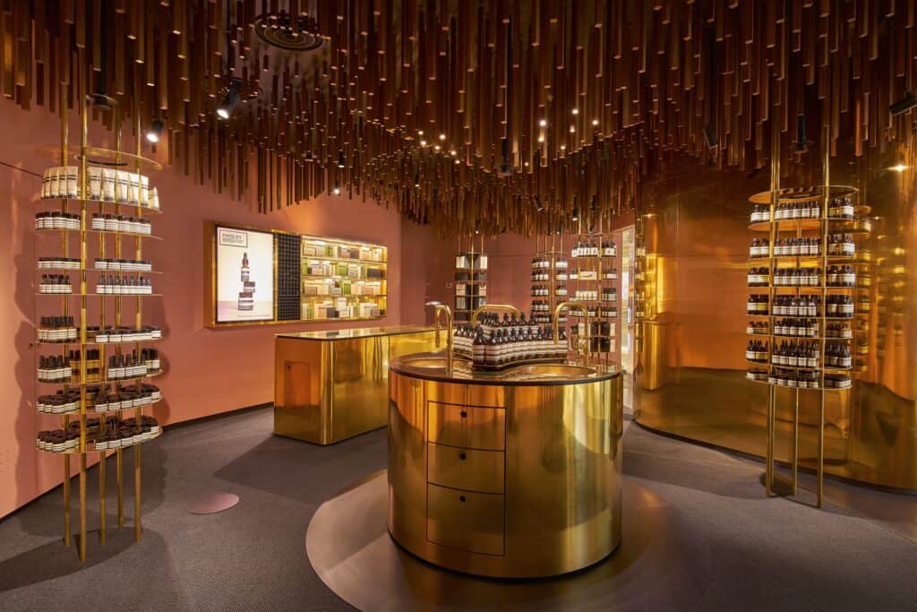 Interior of Aesop ION with a round brass sink on a brass plinth, circular brass shelf towers and a canopy of suspended timber battens over dark sisal carpet