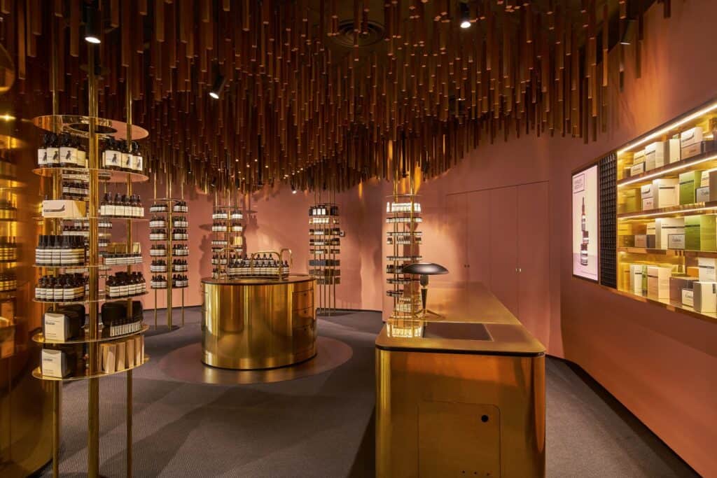 Brass till counter with a black dome lamp beside a backlit wall cabinet of boxed products at Aesop ION, the round brass sink and shelf towers beyond