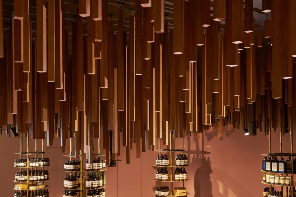 Close-up of square timber battens of varying lengths hanging in dense clusters above brass shelf towers and a pink wall at Aesop ION
