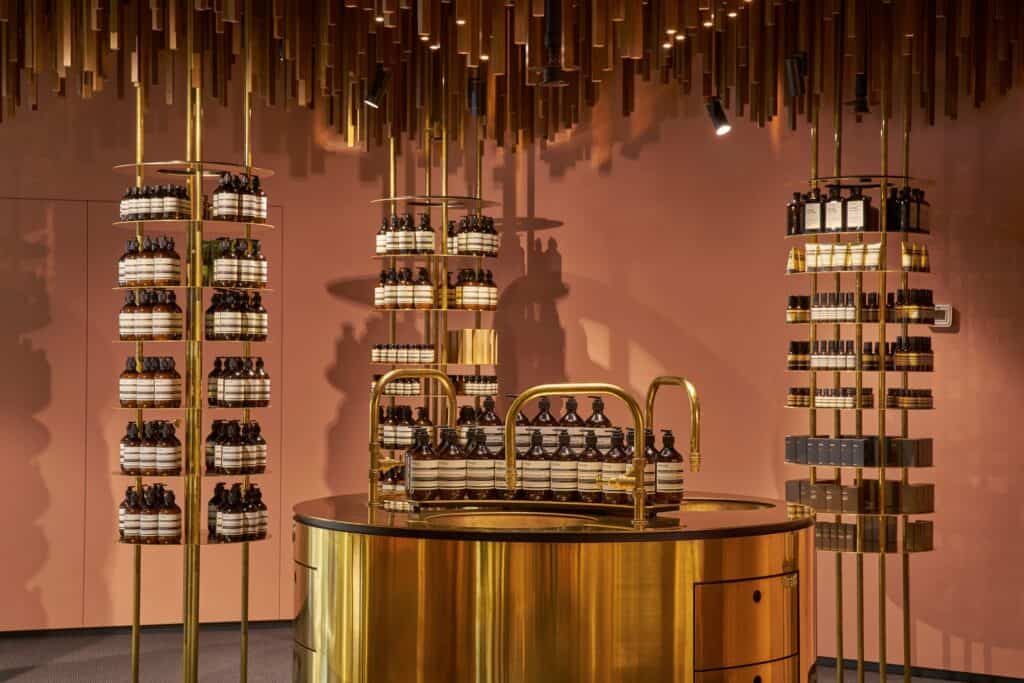Brass swan-neck taps over a round brass basin of hand wash bottles, framed by tiered brass shelf towers against a mace-pink wall at Aesop ION