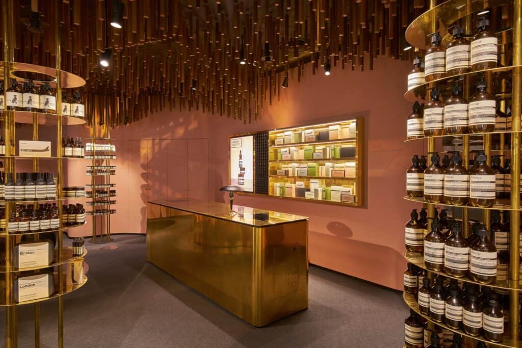 Rectangular brass counter facing a lit brass-framed product cabinet in the mace-pink interior of Aesop ION, a tower of amber bottles in the foreground