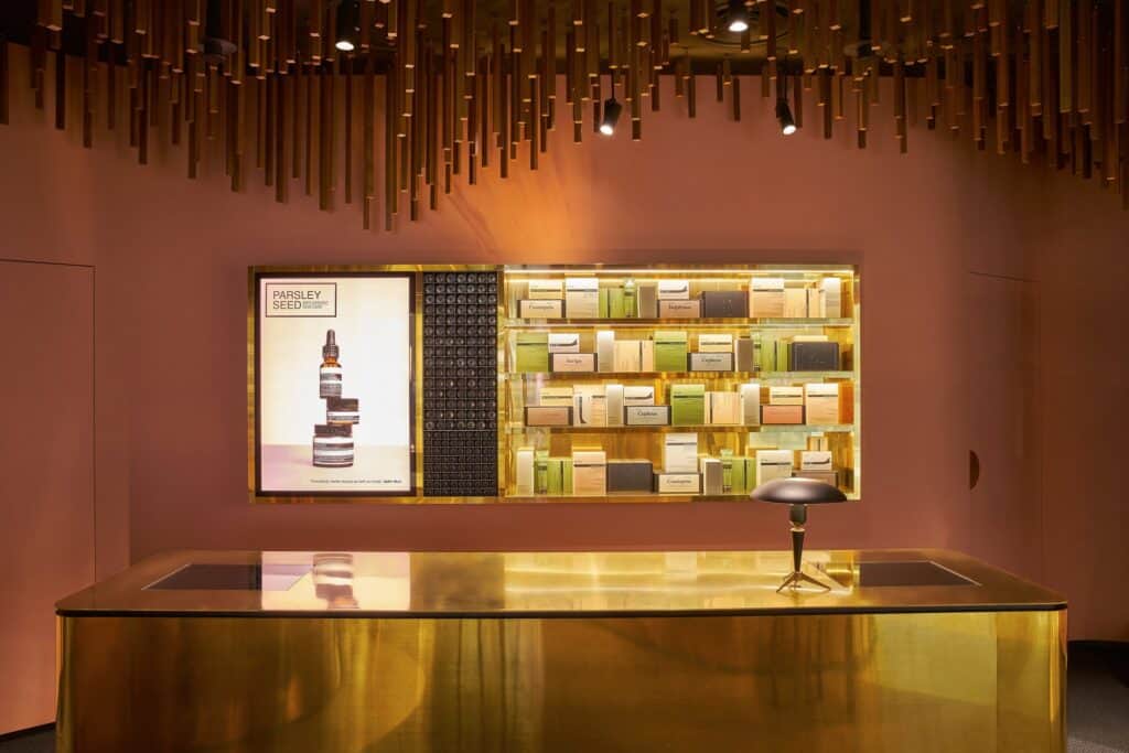 Brass sales counter with a black mushroom lamp before a backlit brass display of boxed Aesop products and a Parsley Seed lightbox at Aesop ION