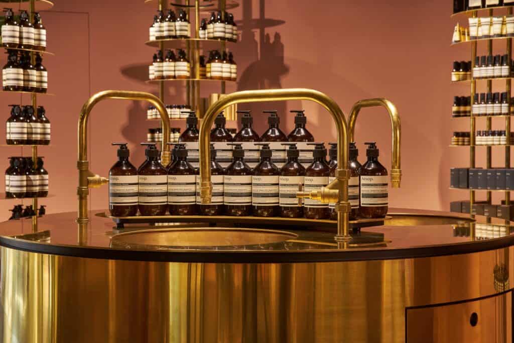 Close-up of brass swan-neck taps and rows of amber Aesop hand wash bottles on the polished rim of a cylindrical brass sink