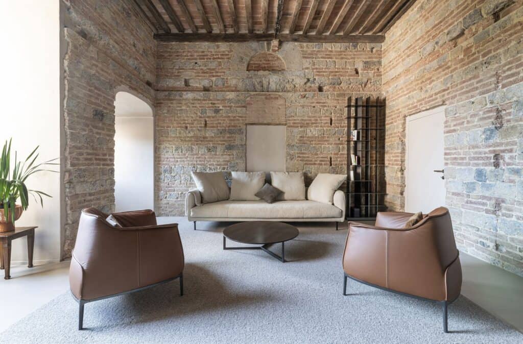 Living room inside the medieval tower of Palazzo Rinuccini in Siena, two leather armchairs and a pale sofa around a round coffee table on a grey rug, framed by restored walls of grey stone and red brick under an elm-beamed ceiling