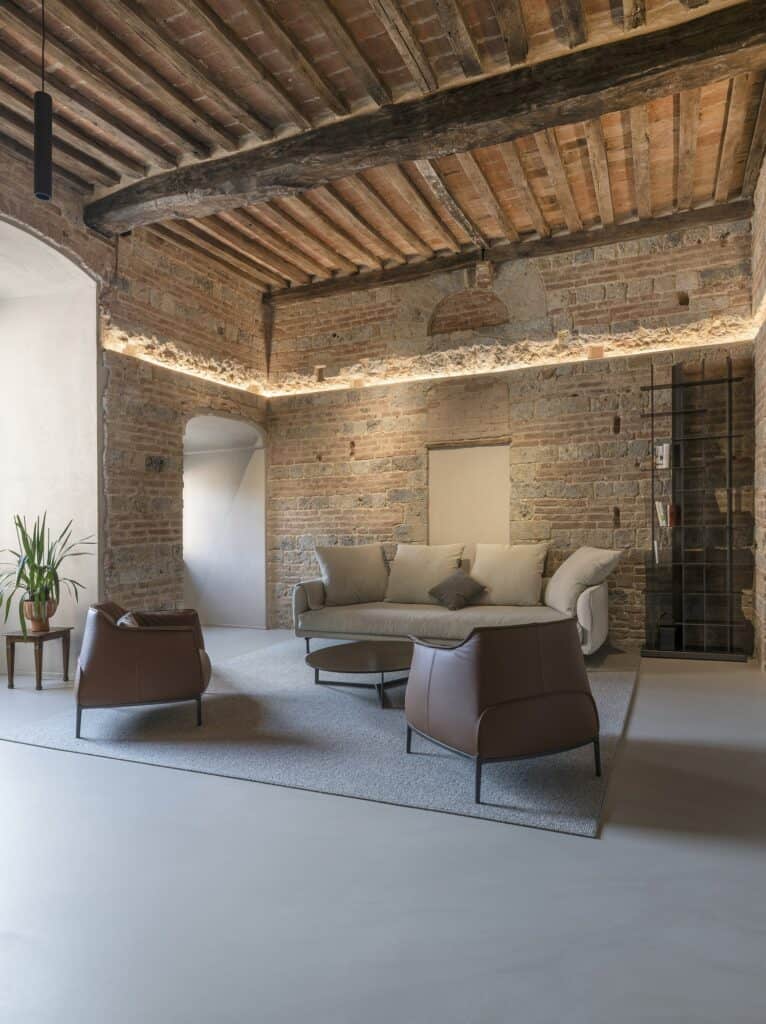 Sitting area of the Apartment inside a Medieval Tower with concealed light washing the top of the brick and stone walls beneath a dark beamed ceiling, an arched doorway, leather armchairs, a sofa and a tall black metal bookcase