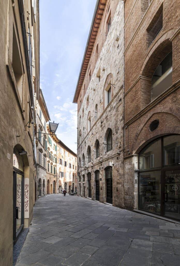 Narrow paved street in the historic centre of Siena lined with medieval palazzi of grey stone and red brick, rows of arched windows and a wrought-iron lantern under a pale blue sky