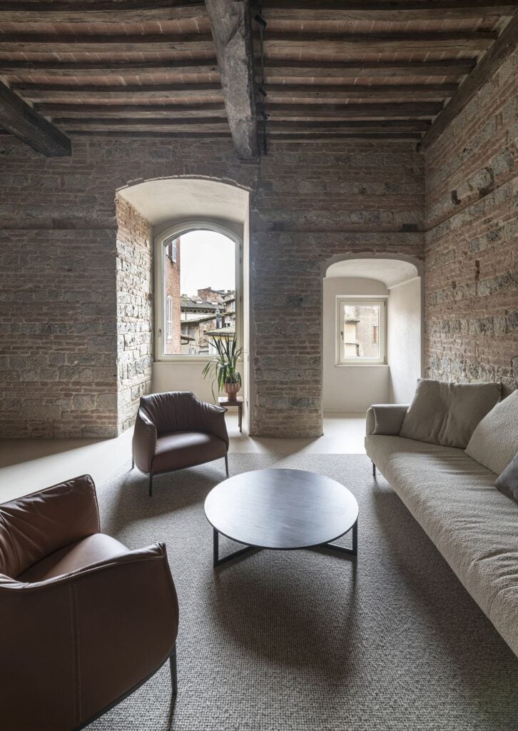 Living room in the medieval tower looking towards two deep arched window openings in the thick stone and brick walls, with leather armchairs, a long pale sofa and a round dark coffee table beneath timber beams