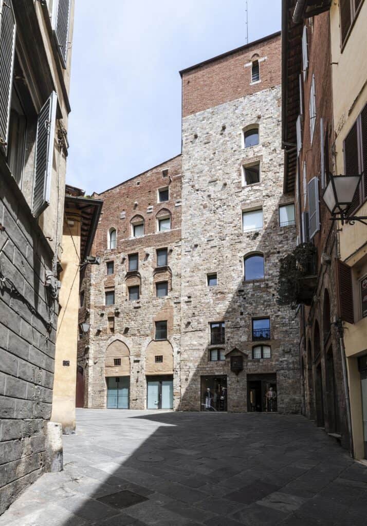 Tall medieval tower-house of grey stone with small arched windows rising above a shaded paved square in Siena, flanked by brick palazzi and a wall lantern