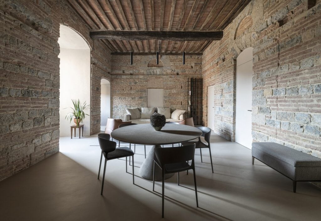 Dining area of the Apartment inside a Medieval Tower with a round pale table, dark chairs and a long upholstered bench on a light resin floor, the living room beyond between restored stone and brick walls