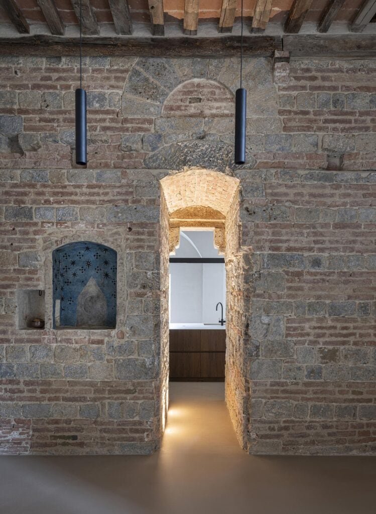 Restored two-tone wall of stone and brick with a blind arch, a blue star-painted niche and a small recess, beside a brick-vaulted doorway lit from below that leads to the kitchen, two slim black pendants hanging above
