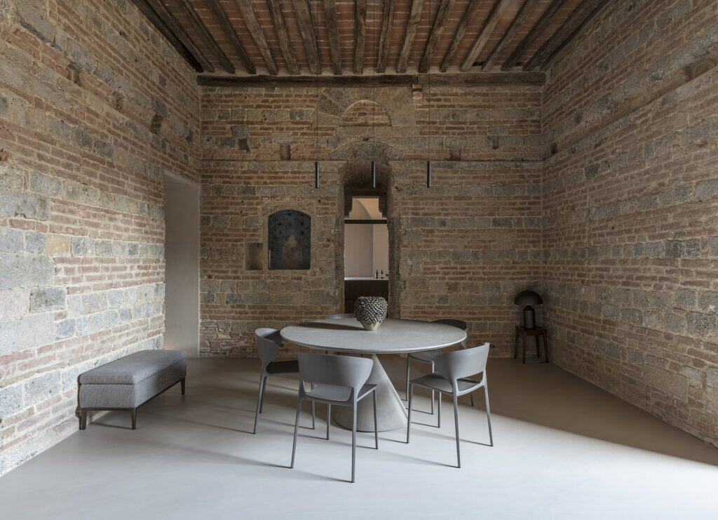 Frontal view of the dining area in the medieval tower, a round table with grey chairs and a grey bench against walls of banded stone and brick beneath a timber and terracotta ceiling, an arched opening to the kitchen at the centre