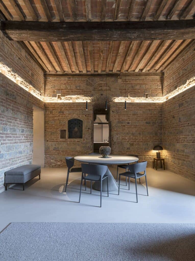 Dining area in the medieval tower with hidden lighting glowing along the top of the stone and brick walls under a heavy timber beam, a round table and grey chairs, a small lamp on a stool and a grey rug in the foreground