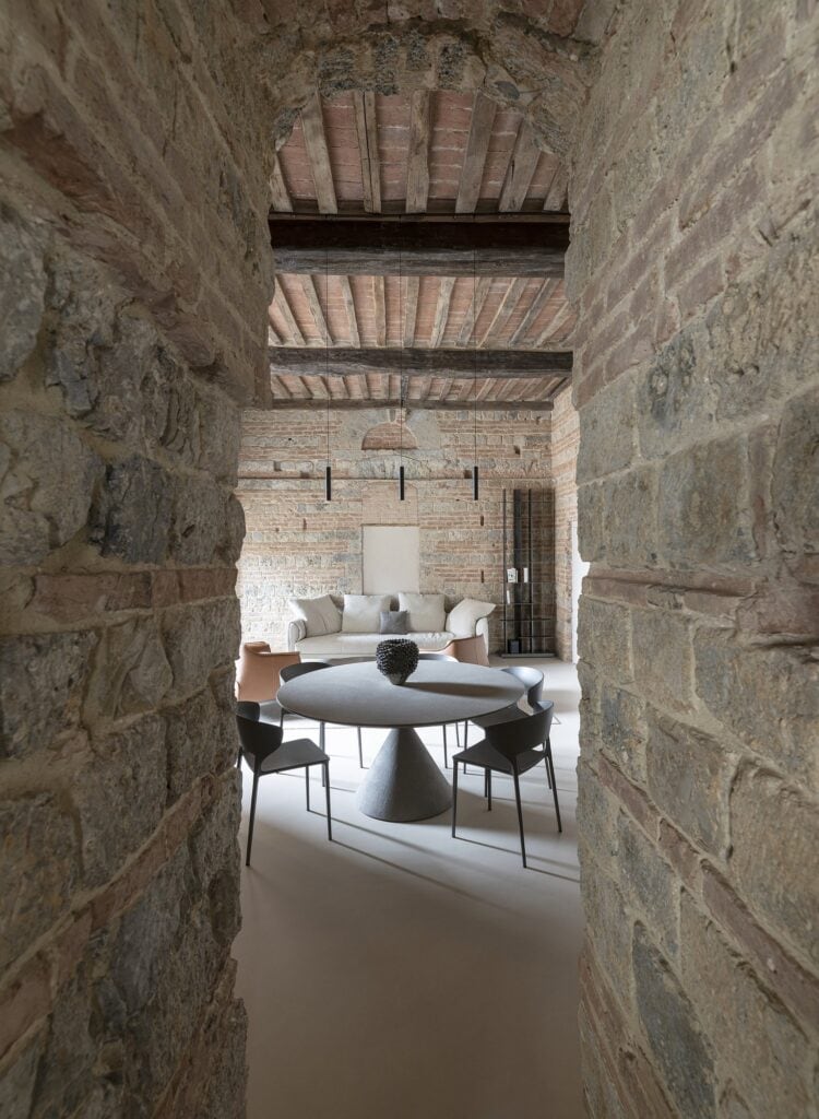 View through a deep opening in the rough stone wall of the tower into the dining and living room, a round table and chairs in front of a pale sofa under a beamed ceiling