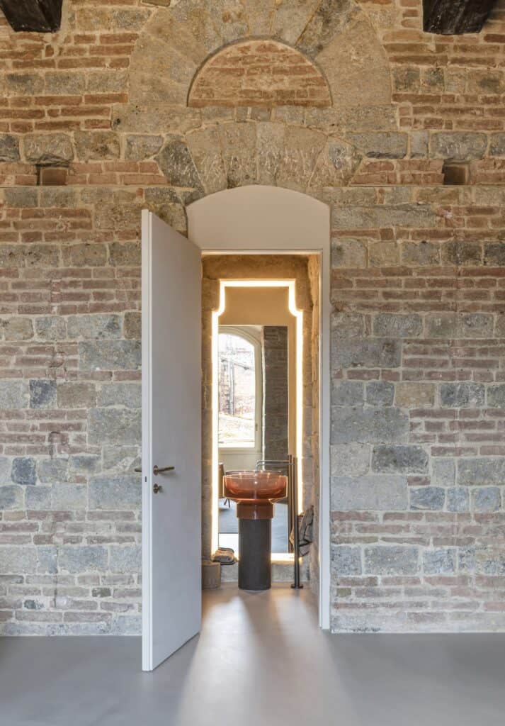White flush door open in a stone and brick wall onto a guest bathroom carved into the tower masonry, an orange glass basin on a dark cork pedestal before an illuminated mirror set into a former monophore window
