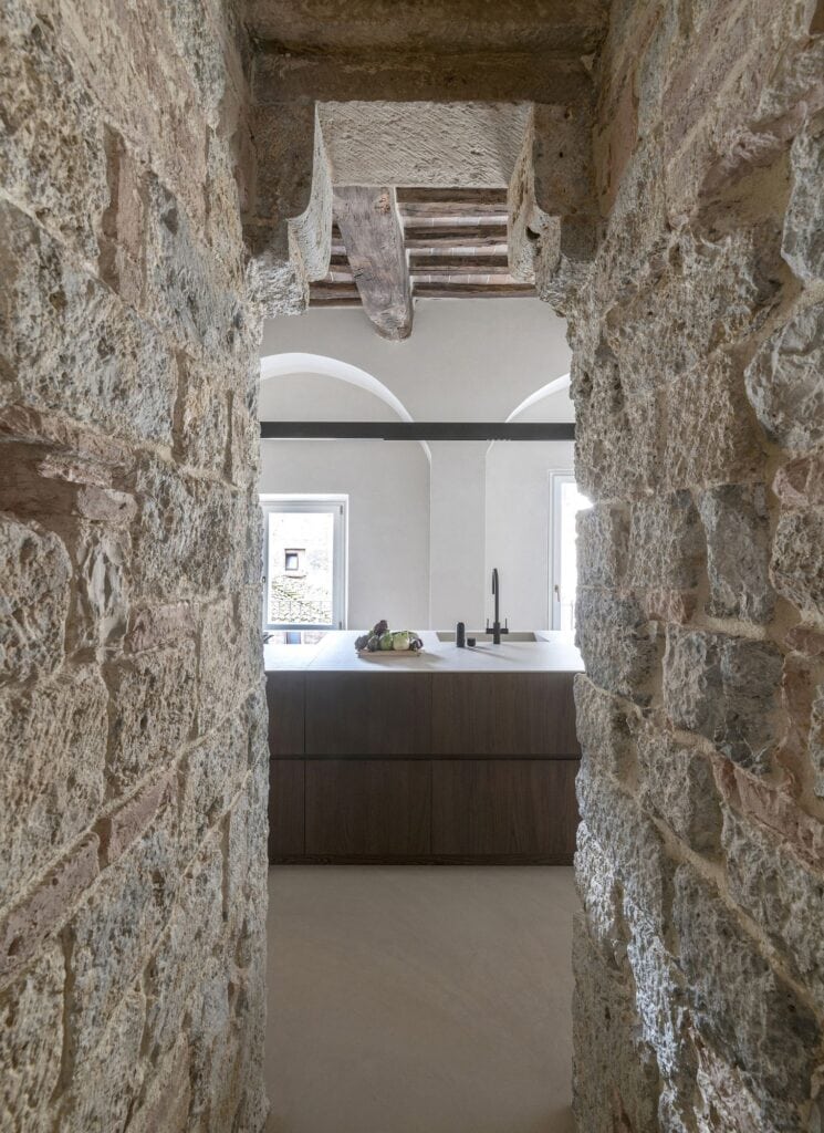 Narrow passage through the rough stone of the medieval tower wall framing the kitchen beyond, a dark wood island and black tap before white arched walls and a window