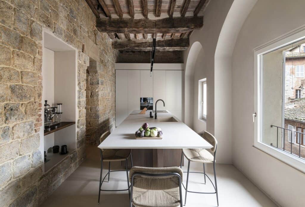 Kitchen beside the medieval tower with a long island of beige ceramic and dark elm, woven-seat bar stools, an old stone wall with a lit shelf niche on the left and a large window and white arches on the right beneath timber beams