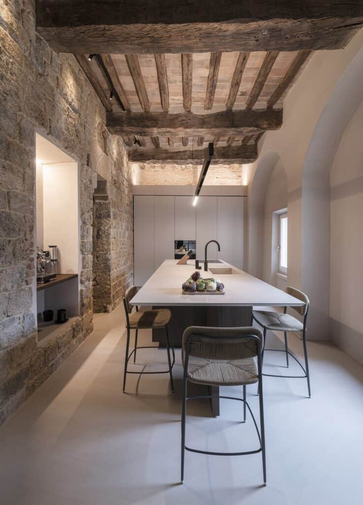 Kitchen island with a beige ceramic top and woven stools running beneath a medieval ceiling of elm beams and terracotta tiles, a black pendant light above and tall pale cabinets at the far end