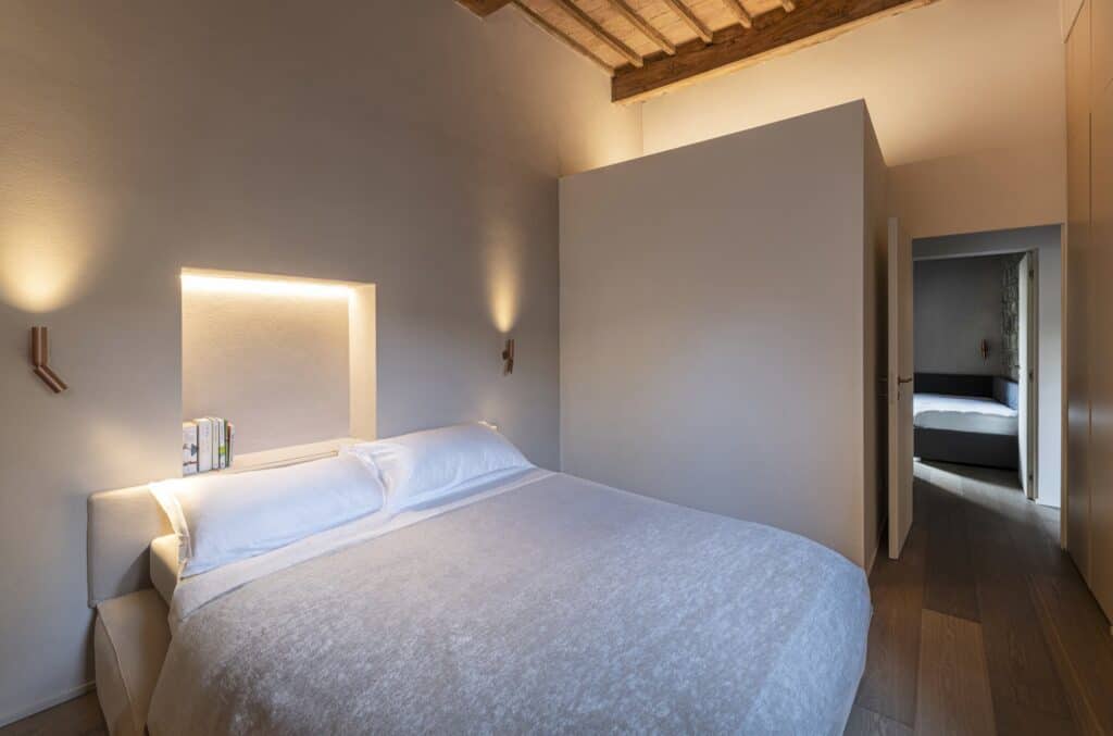 Master bedroom with a double bed beneath a softly lit wall niche, two slim copper wall lights, lime-finished walls, an oak plank floor and a beamed ceiling, a doorway leading through to the second bedroom