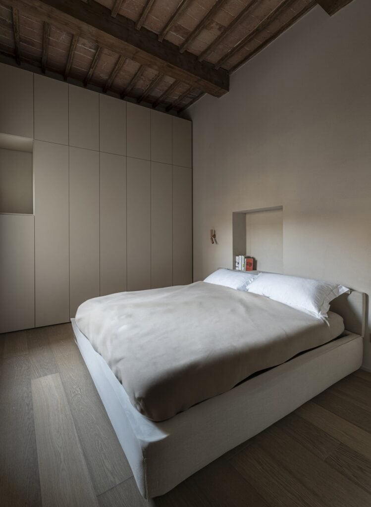 Double bed with white linen before a wall of full-height pale wardrobes, a small lit niche above the bed, an oak plank floor and a sloping ceiling of dark timber beams and terracotta tiles