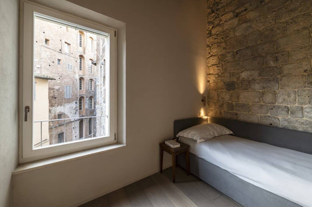 Single bedroom with a grey upholstered bed beside an exposed medieval stone wall lit by a wall lamp, a small side table and a large window looking out onto stone and brick palazzi