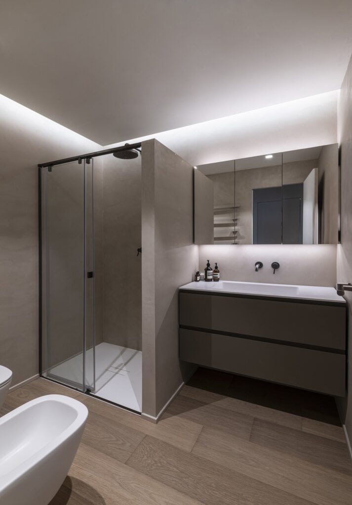Bathroom with a glass shower enclosure, a dark wood vanity with a white basin and black wall-mounted tap, a mirrored cabinet and a white bidet, beneath a ceiling lit along its edges above an oak floor