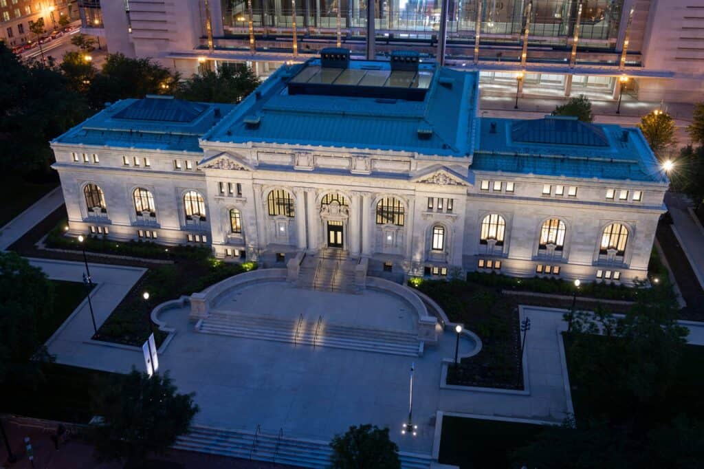 Aerial view of Apple Carnegie Library at dusk, the white marble Beaux-Arts library with lit arched windows and a blue-green roof rising behind a curved granite plaza and steps on Mount Vernon Square