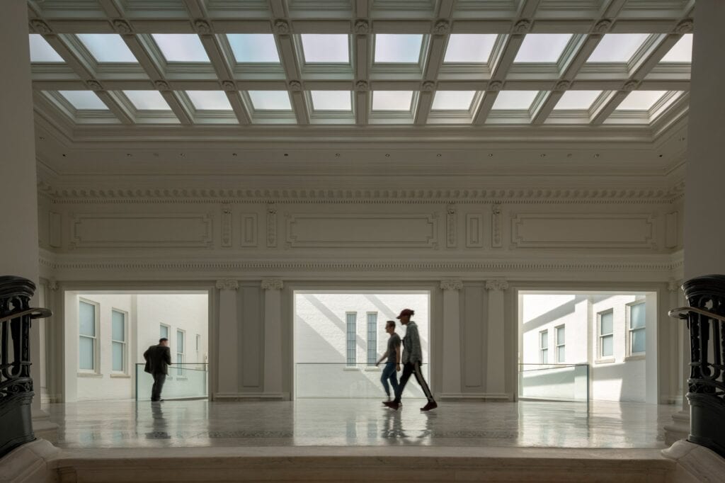 Restored skylit hall at Apple Carnegie Library, a coffered glass ceiling above a white classical frieze and panelled walls, visitors silhouetted walking past glass balustrades framed by dark iron stair railings