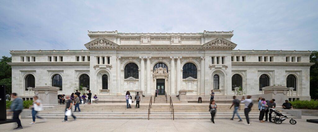 Panoramic view of the south facade of Apple Carnegie Library, the restored white marble library with columns, arched windows and carved pediments above broad granite steps busy with visitors