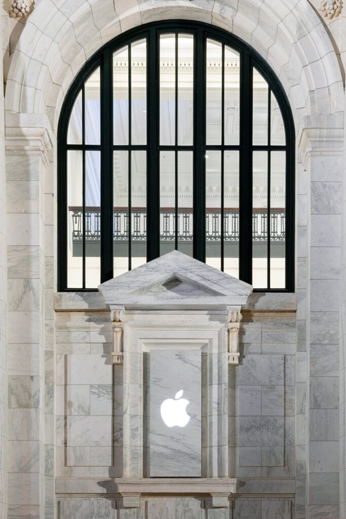 White Apple logo set in a pedimented marble aedicule at Apple Carnegie Library, beneath a tall arched black-framed window looking onto an ornamental balcony railing inside