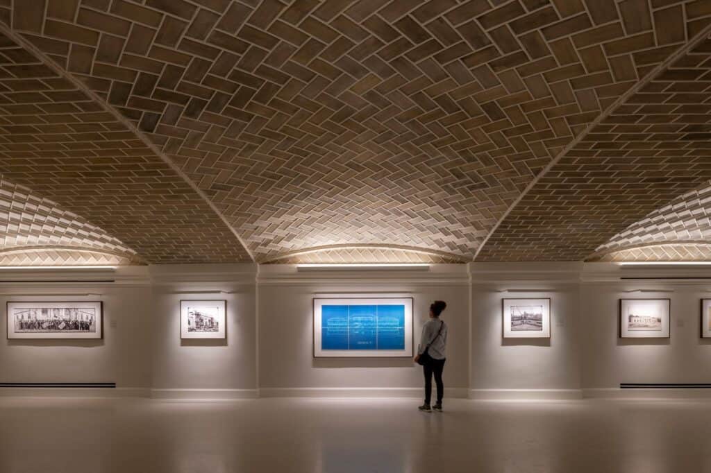 Guastavino vaulted ceiling of herringbone terracotta tiles over the basement gallery at Apple Carnegie Library, a visitor looking at a blue architectural drawing among framed historic photographs