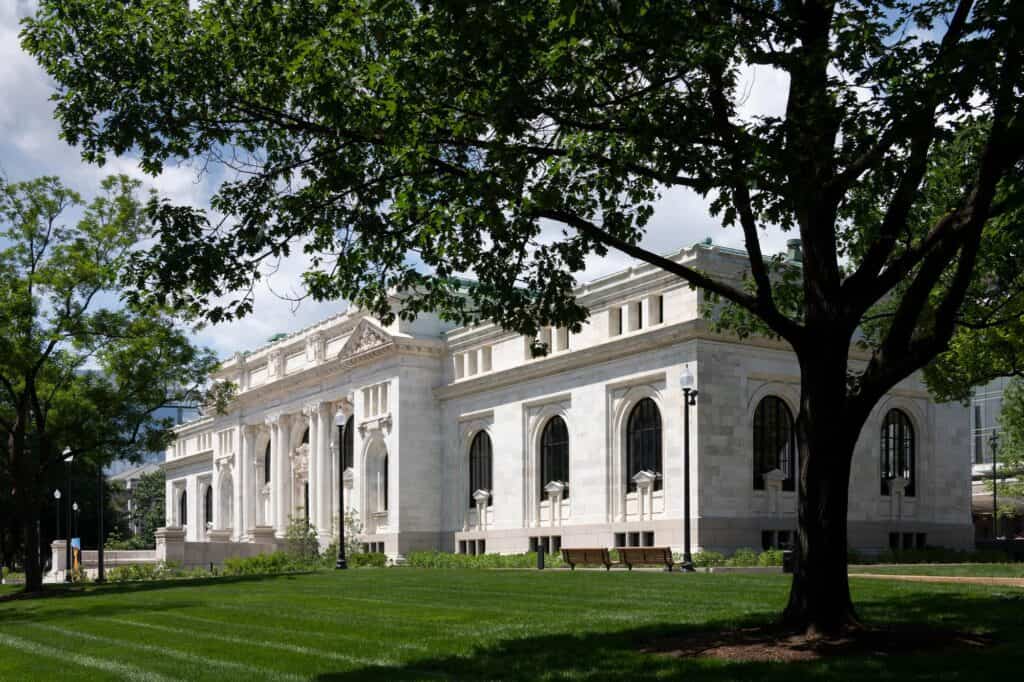 Side view of the restored marble facade of Apple Carnegie Library behind a large shade tree, arched windows and a carved pediment above a green lawn with benches