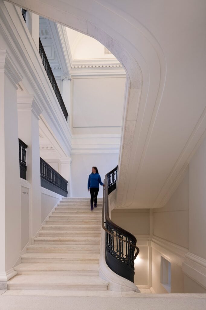 Restored white marble grand staircase at Apple Carnegie Library with a curving black ornamental iron balustrade, a visitor descending beneath a sweeping arch and classical pilasters