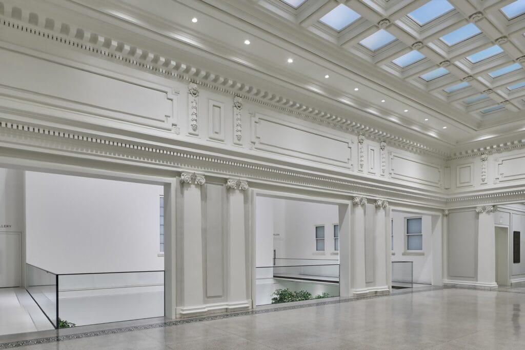 Empty second-floor hall at Apple Carnegie Library beneath a coffered skylight, restored plaster cornices, Ionic pilasters and moulded panels around openings with glass balustrades over the atrium