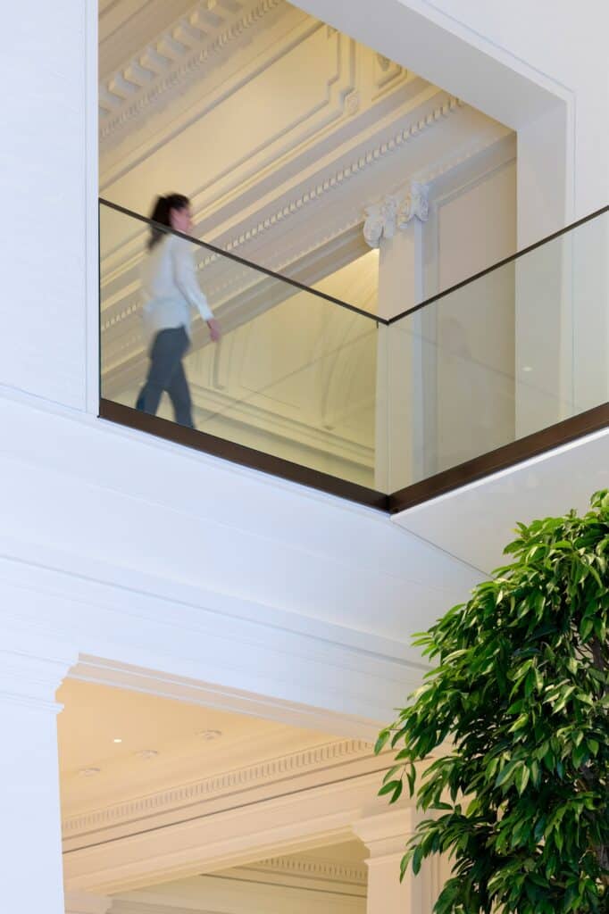 Visitor walking along a glass-balustraded balcony at Apple Carnegie Library past carved classical plasterwork, a ficus tree in the foreground below