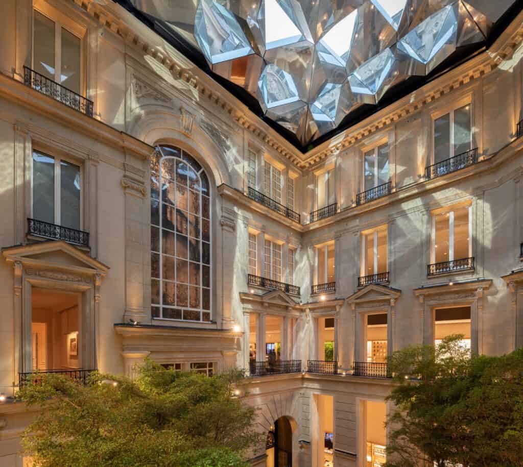 Courtyard of Apple Champs-Élysées at dusk, restored stone facades with a tall arched leaded window and wrought-iron balconies rising to the mirrored pyramids of the Kaléidoscope roof-light, trees below
