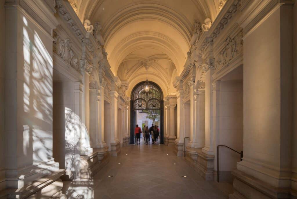Restored 19th-century entrance passage of Apple Champs-Élysées, a carved barrel-vaulted ceiling and stone pilasters leading to an arched wrought-iron gate with a lantern and visitors walking in