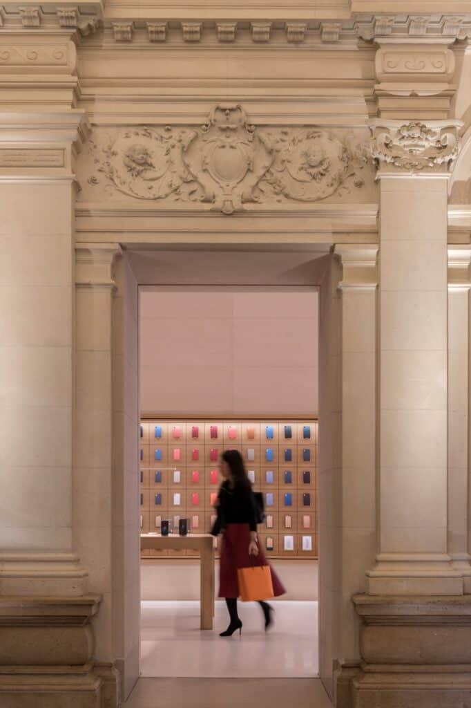 A woman with an orange Apple bag walking through a carved stone doorway with a cherub frieze into a room lined with colourful iPhone cases at Apple Champs-Élysées