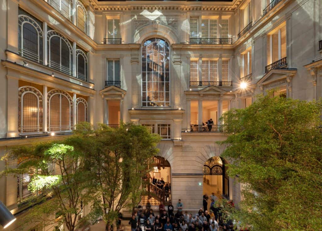 Crowd gathered in the tree-filled courtyard of Apple Champs-Élysées beneath lit stone facades and arched windows, the timber grand staircase visible through an arched opening