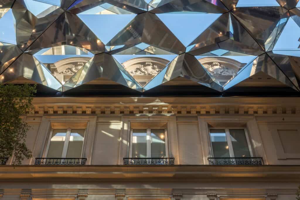 Mirrored pyramids of the Kaléidoscope roof-light at Apple Champs-Élysées reflecting carved window pediments, above a stone cornice and windows with iron balconies