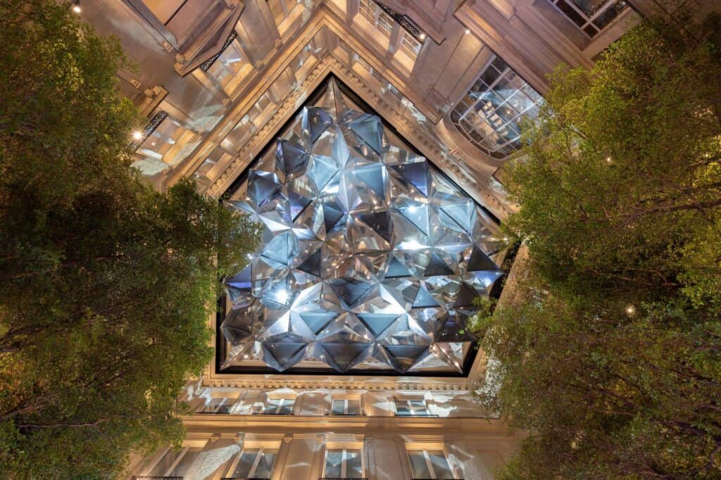 View straight up from the courtyard of Apple Champs-Élysées through tree canopies to the pentagonal Kaléidoscope roof-light framed by illuminated stone facades
