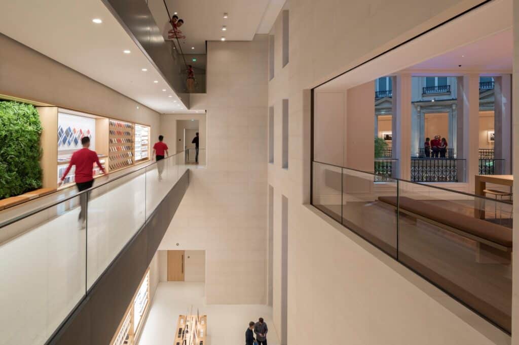 Upper-floor gallery at Apple Champs-Élysées, a glass balustrade beside a green wall and accessory displays overlooking a multi-level pale stone atrium, with a window onto the courtyard