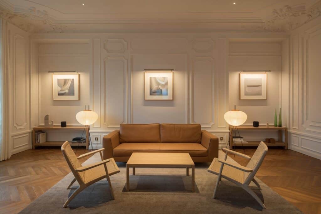 Restored apartment room at Apple Champs-Élysées with panelled walls, ornate plaster cornices and herringbone oak parquet, a tan leather sofa, cane armchairs and paper floor lamps
