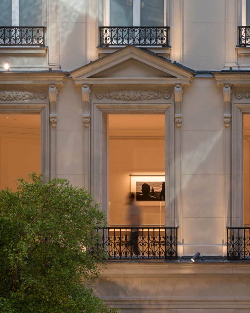 Lit window on the courtyard facade of Apple Champs-Élysées, a carved stone pediment and wrought-iron balcony framing a black-and-white photograph inside, beside a tree